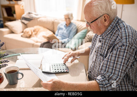 Vista laterale ritratto di uomo senior il riempimento di forme e di pagare le tasse mentre è seduto a tavola con donna anziana e cane seduti sul divano in background, Foto Stock