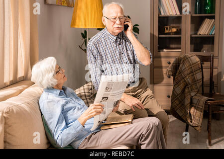 Ritratto della moderna coppia senior di parlare al telefono e leggere giornali seduto sul divano nel soleggiato soggiorno, spazio di copia Foto Stock