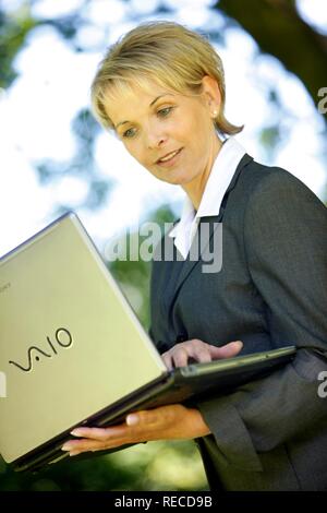 Donna che indossa un ladies suit, pantaloni adatti, imprenditrice, inizio 40s, lavorando sul suo computer portatile in un parco Foto Stock