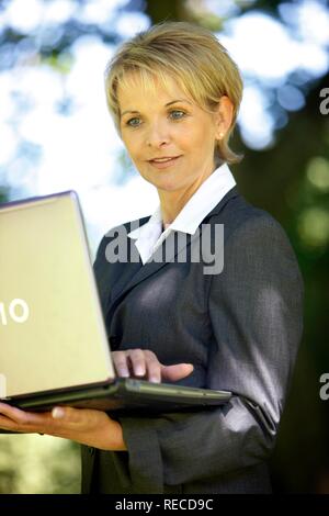 Donna che indossa un ladies suit, pantaloni adatti, imprenditrice, inizio 40s, lavorando sul suo computer portatile in un parco Foto Stock