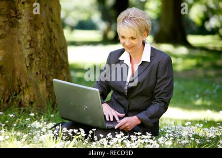 Donna che indossa un ladies suit, pantaloni adatti, imprenditrice, inizio 40s, lavorando sul suo computer portatile in un parco Foto Stock