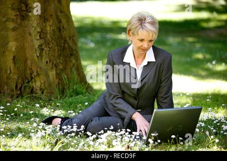 Donna che indossa un ladies suit, pantaloni adatti, imprenditrice, inizio 40s, lavorando sul suo computer portatile in un parco Foto Stock