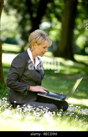 Donna che indossa un ladies suit, pantaloni adatti, imprenditrice, inizio 40s, lavorando sul suo computer portatile in un parco Foto Stock