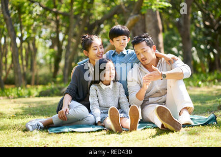 Famiglia asiatica con due bambini divertirsi seduti su erba a parlare in chat all'aperto nel parco Foto Stock