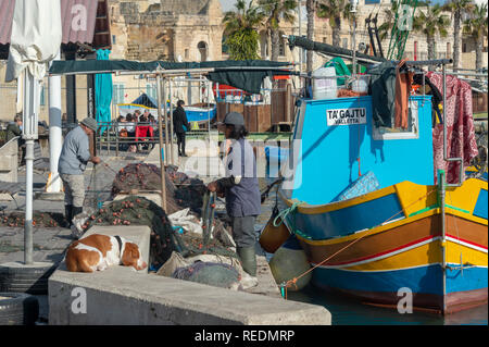 I pescatori a riparare le reti quayside di Marsaxlokk, Malta accanto a loro tradizionale barca da pesca. Foto Stock