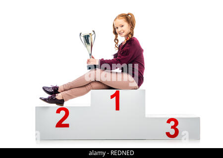 Felice schoolgirl seduti sul vincitore podio, tenendo trophy cup e guardando la telecamera isolato su bianco Foto Stock