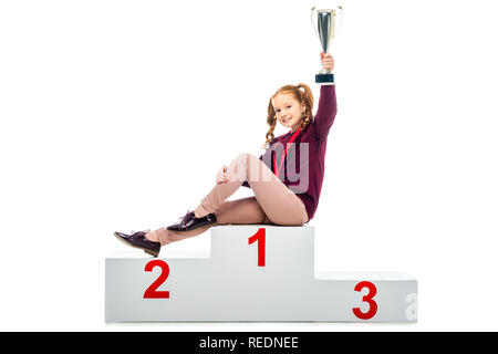 Sorridente schoolgirl seduti sul vincitore podio, tenendo trophy coppa in mano sollevata e guardando la telecamera isolato su bianco Foto Stock