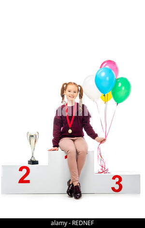 Sorridente schoolgirl seduto sul podio vincitore trofeo vicino a tazza, tenendo palloncini colorati e guardando la telecamera isolato su bianco Foto Stock