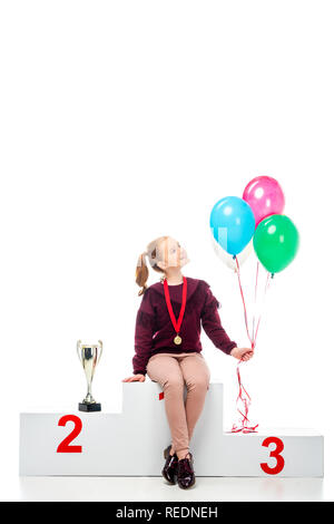 Sorridente schoolgirl seduto sul podio vincitore trofeo vicino a tazza e azienda palloncini colorati isolato su bianco Foto Stock