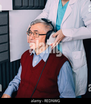 Senior paziente che indossa le cuffie, seduto in cabina insonorizzata, durante l'esame dell'orecchio a audiologi office Foto Stock