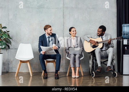 Sorridente imprenditori multiculturale di seduta e di ascolto durante il african american uomo in carrozzella a suonare la chitarra in sala di attesa Foto Stock