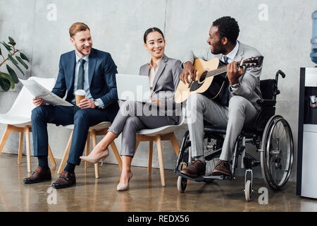 Imprenditori multiculturale di seduta e di ascolto durante il disabilitato african american uomo a suonare la chitarra in sala di attesa Foto Stock