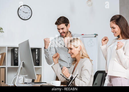 Eccitato mentor di business con i colleghi agitando i pugni e guardando il desktop computer in ufficio Foto Stock