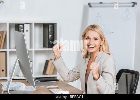 Emozionato imprenditrice agitando i pugni e sorridente alla fotocamera mentre è seduto al posto di lavoro Foto Stock
