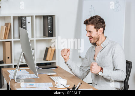 Eccitato giovane impreditore agitando i pugni e guardando il desktop computer in ufficio Foto Stock