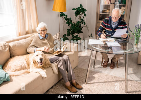Angolo alto ritratto di adorabili golden retriever cane seduto sul divano con la coppia senior a casa illuminata dalla luce del sole, spazio di copia Foto Stock