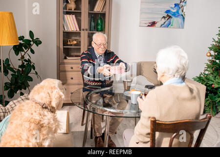 Ritratto di felice coppia senior scambiando presenta seduta tutta la tabella a casa, focus su sorridente uomo vecchio azienda confezione regalo Foto Stock