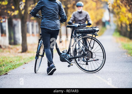 Attivo coppia senior con electrobikes in piedi all'aperto su una strada nel parco. Foto Stock