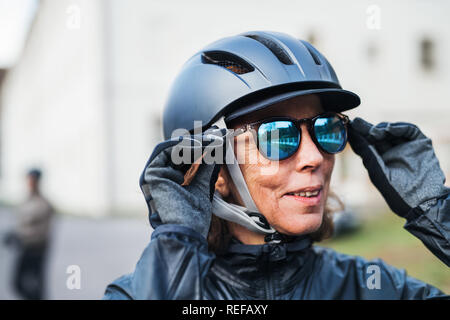 Attiva una donna senior con il casco protettivo in piedi all'aperto, mettendo in occhiali da sole. Foto Stock