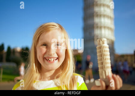 Sorridente ragazza moderna che mostra la torre pendente di Pisa souvenir contro la torre pendente di Pisa, Italia. La ragazza con i capelli biondi in giallo con strisce bianche t-shirt Foto Stock