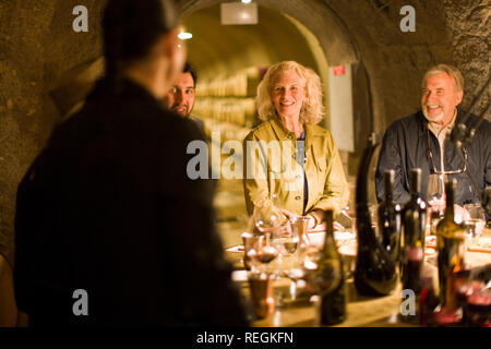 Gruppo di sorridere adulti seduti intorno a una tavola rotonda prevista con le bottiglie di vino all'interno di una stanza buia. Foto Stock