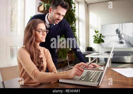 Colpo di giovane e bella segretaria imprenditrice seduto alla scrivania in ufficio e collega in piedi accanto a lei mentre utilizzando laptop e consulenza. Lavoro di squadra Foto Stock