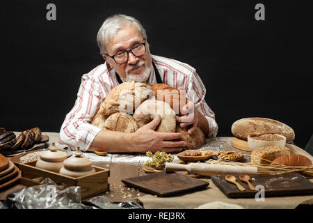 Felice Vecchio baker guardando la fotocamera e sorridere mentre abbraccia le forme di pane Foto Stock