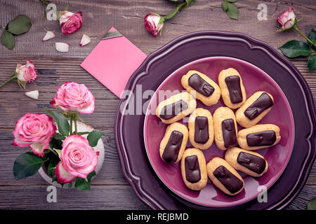 Mini eclairs alla vaniglia con glassa di cioccolato sulla piastra di porpora, vista dall'alto sulla tavola in legno rustico con le rose rosa e la carta regalo Foto Stock