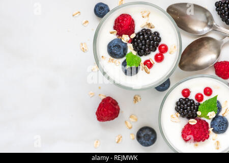 Yogurt sano con freschi frutti di bosco in bicchieri con cucchiai su marmo bianco dello sfondo. Dieta sana prima colazione. vista superiore Foto Stock