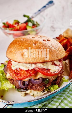 Tradizionale hamburger con tutti i rifili compresi il formaggio fuso, pomodori, lattuga, cipolla e maionese servita in un vecchio piatto vintage con un lato Foto Stock