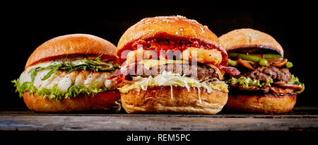 Close-up di tre diversi gustosi hamburger di pesce, carni bovine la carne o il formaggio su un tavolo di legno su sfondo nero per spazio di copia Foto Stock