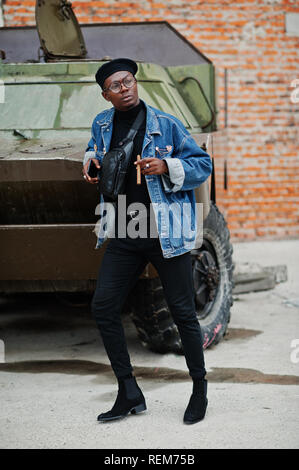 African American Uomo in camicia di jeans, berretto e occhiali, con sigaro poste contro militari btr veicolo blindato. Foto Stock