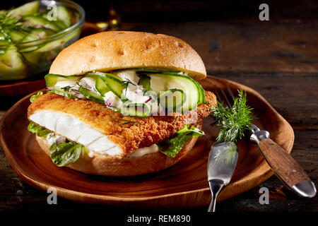 Deliziosi frutti di mare burger con un impanati filetto di pesce, cetriolo e baby spinaci su una piastra con utensili su un rustico sfondo legno Foto Stock