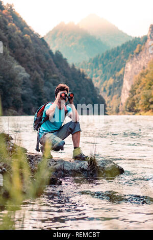 Giovani turisti con zaino guarda attraverso un binocolo sulle cime delle montagne, sorge su una roccia su un fiume. Ragazzo trascorre una vacanza in montagna, wanderin Foto Stock