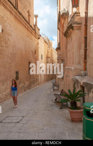 Giovane donna a piedi attraverso le tipiche strade strette tra case di calcare nell'antica città fortificata di Mdina a Malta. Foto Stock