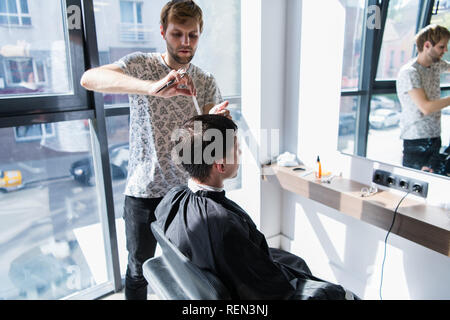 Un parrucchiere professionista con un pettine e forbici in mano lo styling wet nero e capelli corti dell'uomo in un salone di parrucchiere Foto Stock