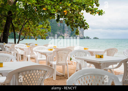 Ao Nang, Tailandia - 2 Luglio 2, 2018: tabelle vuote di un open-air cafè sulla spiaggia Ao Nang contro il cielo nuvoloso e mare grigiastro. Foto Stock