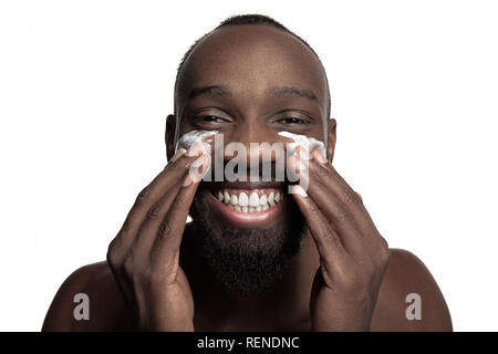 Giovane africano-ragazzo americano l'applicazione di crema per il viso sotto i suoi occhi su sfondo bianco. Ritratto di un giovane sorridenti africano a studio. Alta Moda modello maschile. Foto Stock