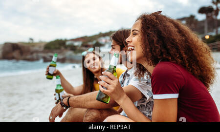Sorridente giovane donna con bottiglia di birra e gli amici seduti da sulla spiaggia. Vista laterale del gruppo di amici avente un party sulla spiaggia di sera. Foto Stock