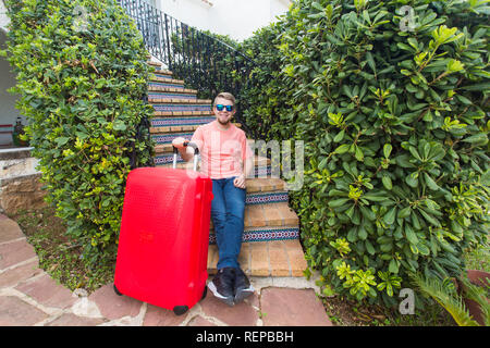 Vacanze, viaggiare e concetto di persone - uomo con red travel valigia seduti sulle scale la sua casa Foto Stock