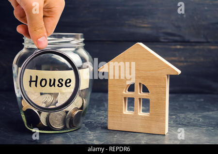 Un vaso di vetro con monete e una casa in legno e la scritta 'casa'. L'accumulo di denaro per comprare una casa. Affitta un appartamento. Acquisto di una casa. Foto Stock