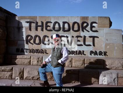 Un uomo che indossa un giubbotto e cappello e tenendo una fotocamera stand con una gamba fino al segno per Theodore Roosevelt National Memorial Park di Medora, North Dakota, 1976. () Foto Stock