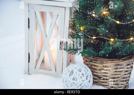 White Lanterna di legno con cera bianca candela accesa e piccole Natale abete con micro luci a led in rattan di colore marrone pentola floreale in snow, innevate di abeti Foto Stock