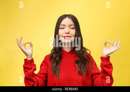 Persone, yoga e uno stile di vita sano. Stupendo giovane donna vestita in rosso chiaro maglione mantenendo gli occhi chiusi mentre medita in ambienti chiusi, praticando la pace della mente, mantenendo le dita nel gesto mudra Foto Stock