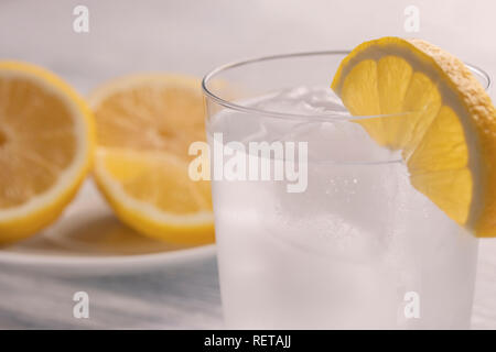 Rinfrescante acqua fredda con limone in un bicchiere bagnato con ghiaccio. Su un bianco tavolo di legno. Pronto per l'uso. Un pezzo di frutta si trova su una piastra bianca. Dimagrimento ref Foto Stock