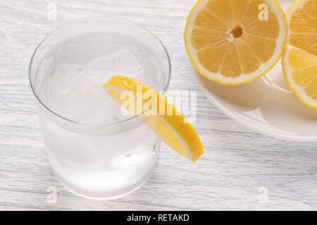 Rinfrescante acqua fredda con limone in un bicchiere bagnato con ghiaccio. Su un bianco tavolo di legno. Pronto per l'uso. Un pezzo di frutta si trova su una piastra bianca. Parte refres Foto Stock