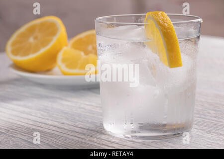 Rinfrescante acqua fredda con limone in un bicchiere bagnato con ghiaccio. Su un bianco tavolo di legno. Pronto per l'uso. Un pezzo di frutta si trova su una piastra bianca. Una sana Drin Foto Stock