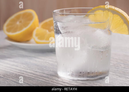 Rinfrescante acqua fredda con limone in un bicchiere bagnato con ghiaccio. Su un bianco tavolo di legno. Pronto per l'uso. Un pezzo di frutta si trova su una piastra bianca. Bevanda utile Foto Stock