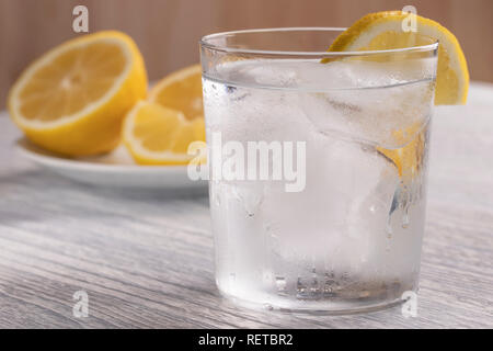 Rinfrescante acqua fredda con limone in un bicchiere bagnato con ghiaccio. Su un bianco tavolo di legno. Pronto per l'uso. Un pezzo di frutta si trova su una piastra bianca. Una sana drin Foto Stock