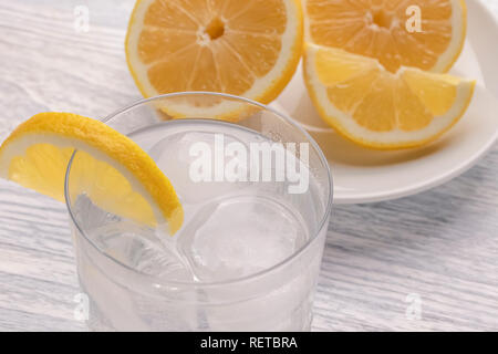 Rinfrescante acqua fredda con limone in un bicchiere bagnato con ghiaccio. Su un bianco tavolo di legno. Pronto per l'uso. Un pezzo di frutta si trova su una piastra bianca. Una sana drin Foto Stock